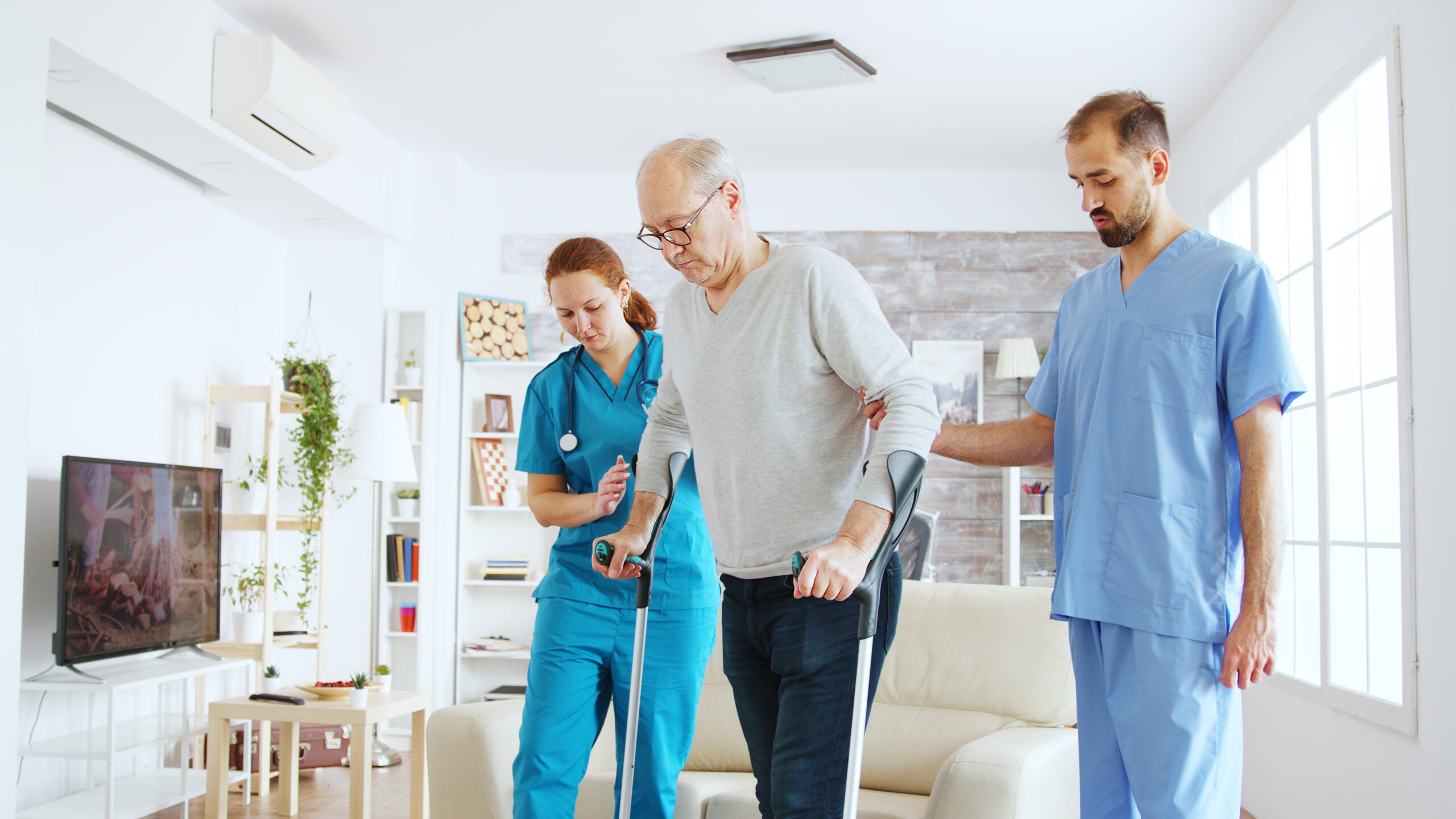 Team of nurses or social workers helping an old disabled man to walk with his crutches out of the nursing home room Team of nurses or social workers helping an old disabled man to walk with his crutches out of the nursing home room